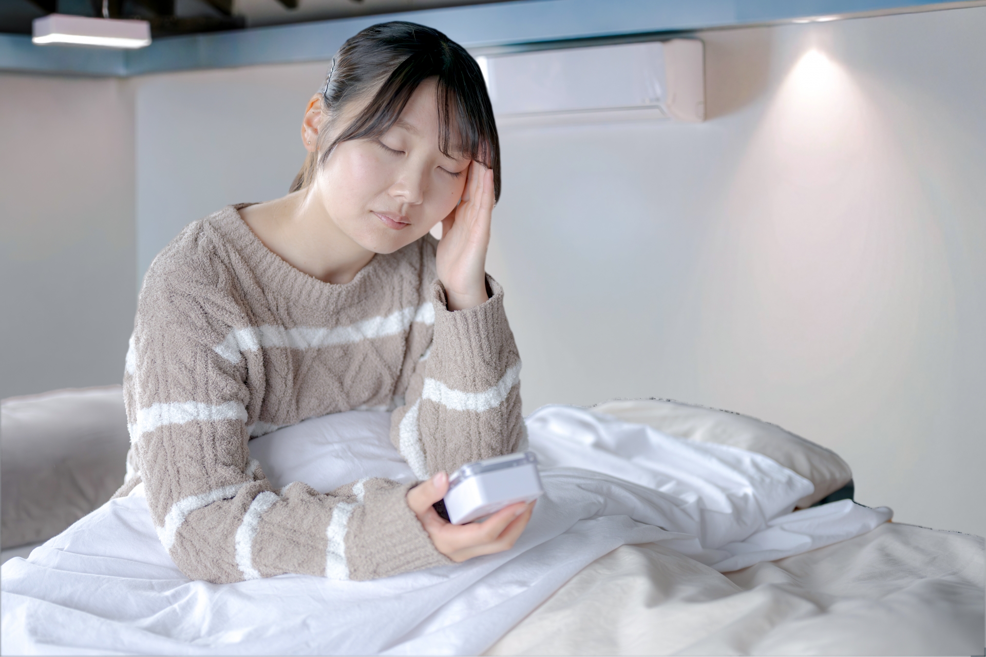 ベッドで眠れずにスマートフォンを持ちながら頭を抱えている女性。不眠・睡眠障害のイメージ