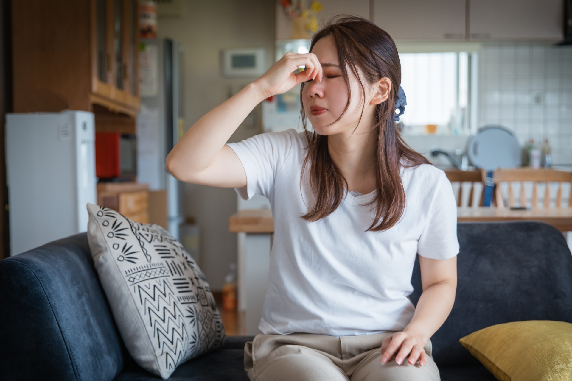 ソファに座り、額に手を当てて目を閉じている女性。めまいや頭の重さでつらそうな表情