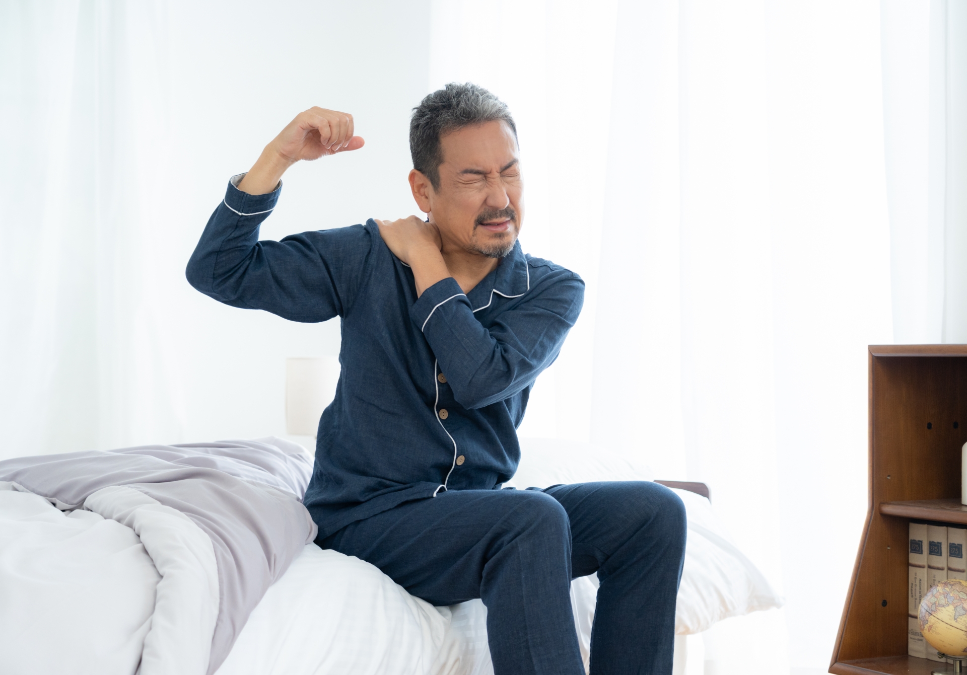 ベッドの上で肩を押さえて痛みに顔をしかめている男性。五十肩・肩関節痛のイメージ