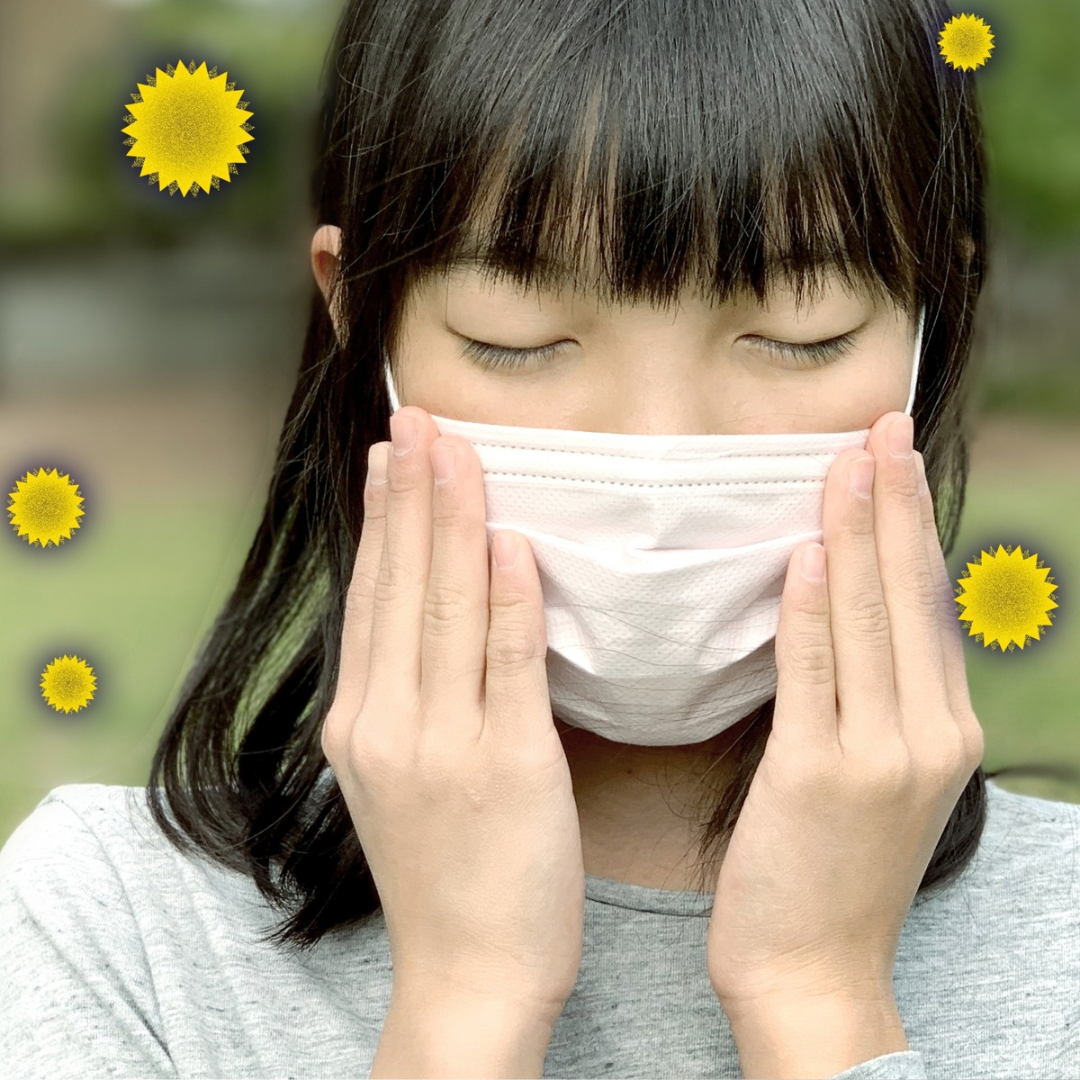 マスクを両手で顔に当てる女性の写真。周囲に花粉を表す黄色い花のアイコンが描かれており、花粉症対策のイメージ。